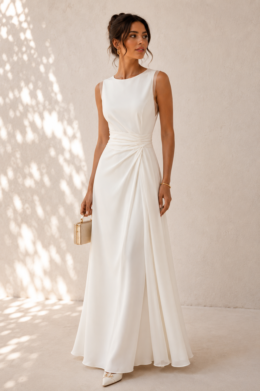 A woman modeling a sleeveless white floor-length gown with a gathered waist and flowing skirt, standing against a textured beige wall with dappled sunlight and shadows.
