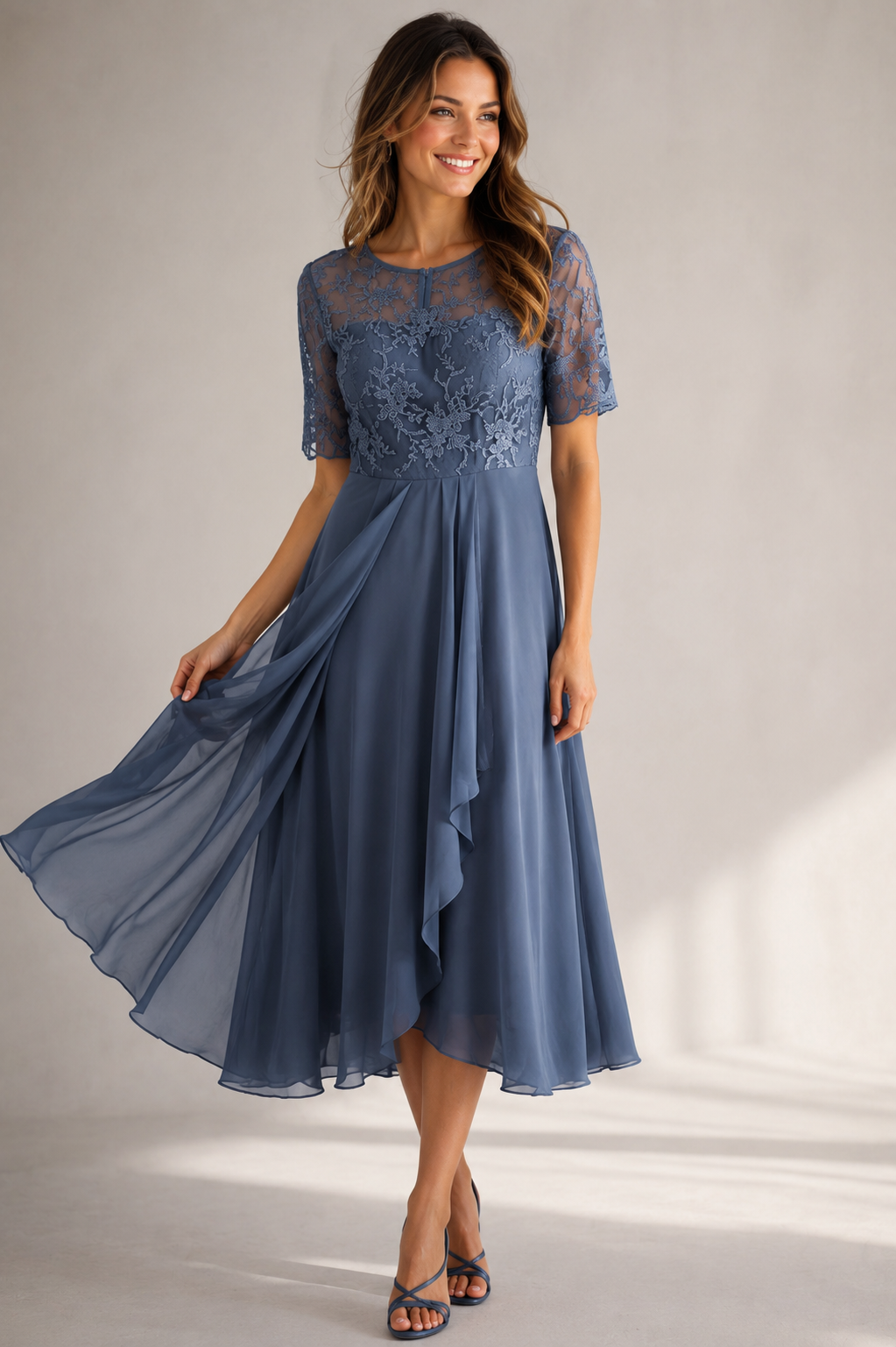 A woman modeling a dusty blue midi dress with a sheer lace bodice, short sleeves, and a flowing chiffon skirt, set against a neutral studio background.
