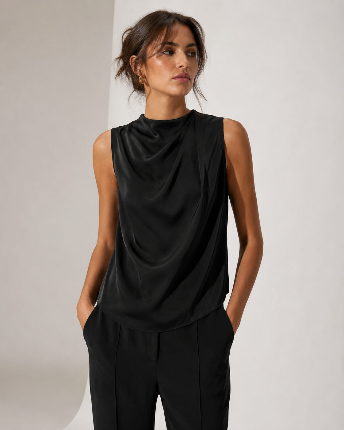 A woman modeling a black sleeveless draped top with a high mock neck, paired with black trousers, against a clean light gray studio background.