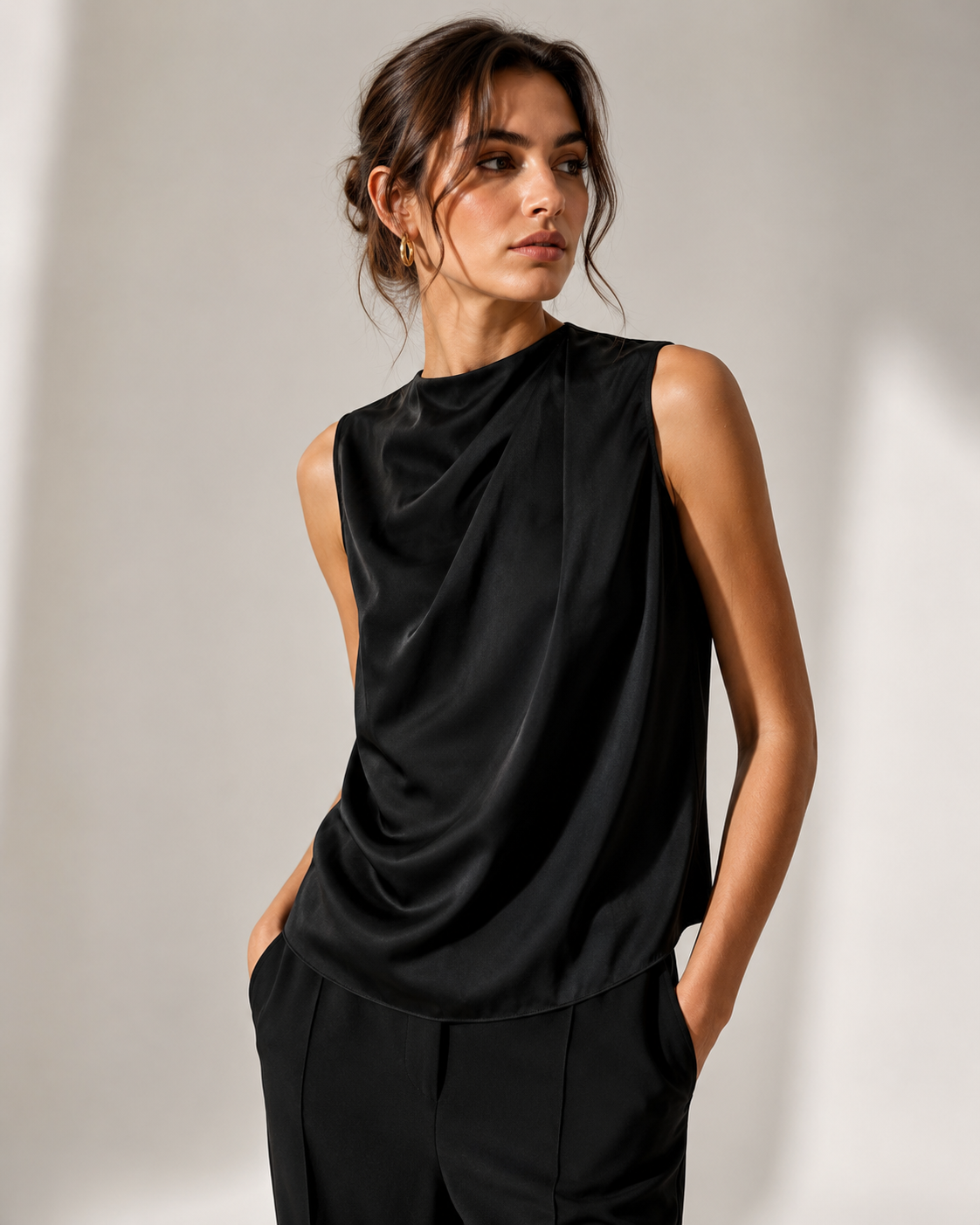 A woman wearing a black sleeveless draped top and matching black trousers, standing against a light gray studio wall with shadows.