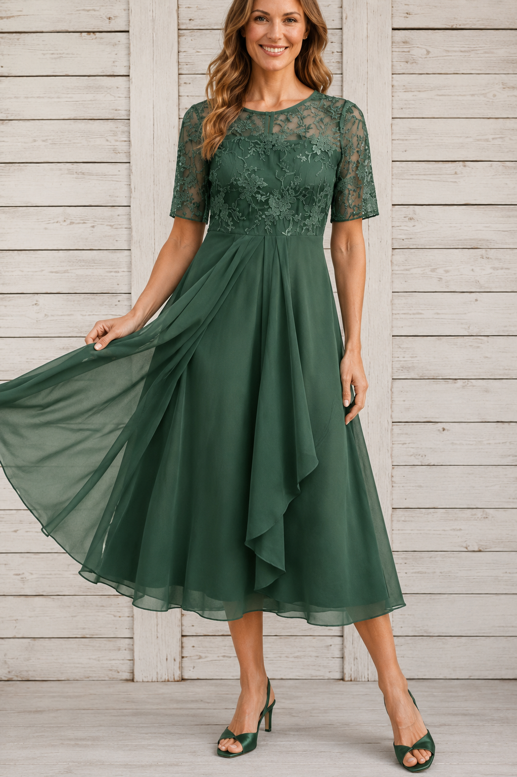 A woman modeling a dark green midi dress with a lace bodice, sheer short sleeves, and a flowing chiffon skirt, standing against a white wooden plank wall.