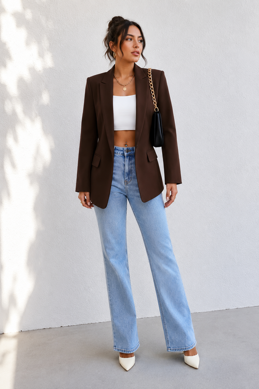 A woman stands against a light gray wall wearing an oversized dark brown blazer over a white cropped tank top, light blue wide-leg jeans, and white pointed heels. She is carrying a black shoulder bag with a gold chain strap.