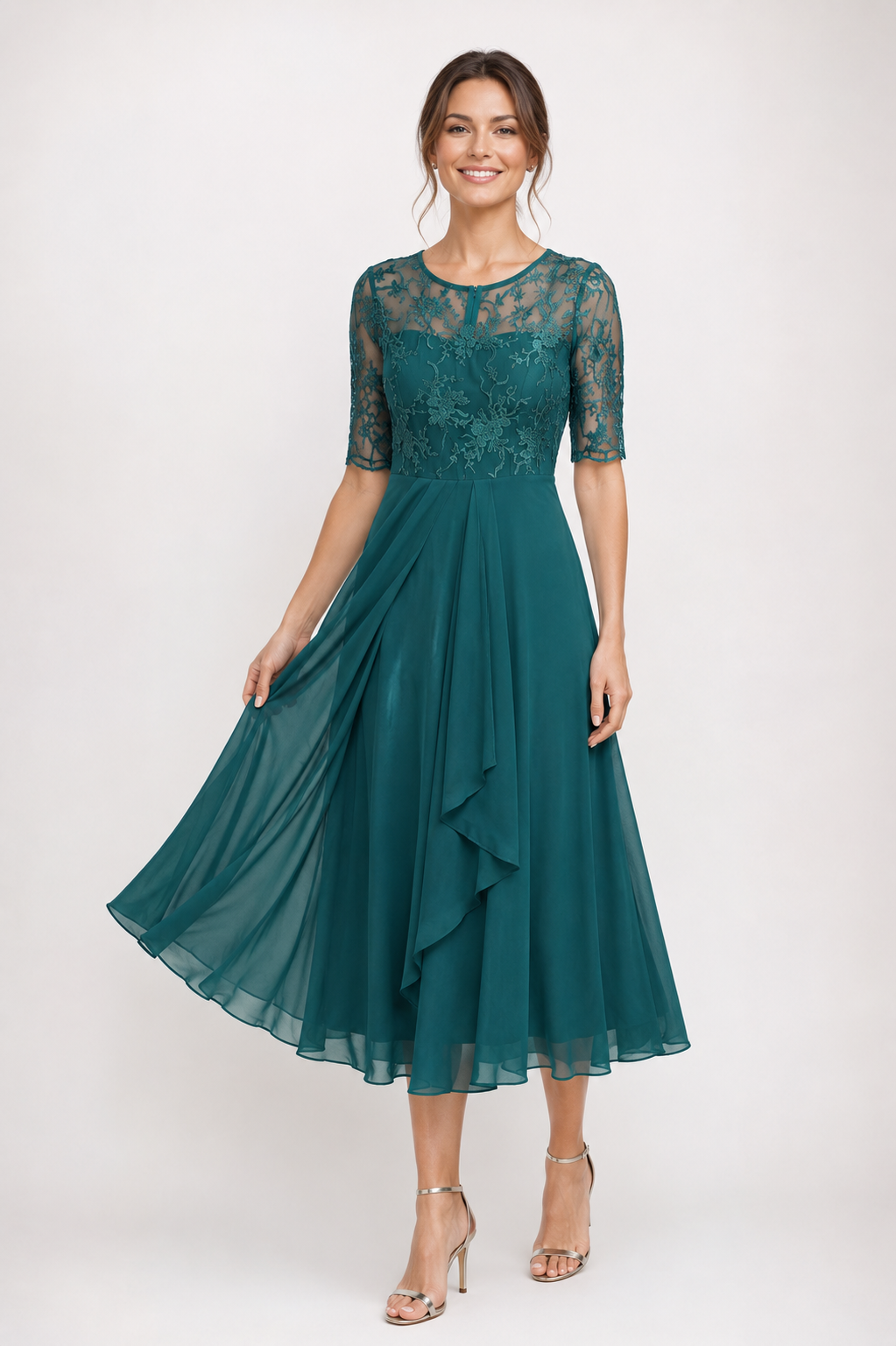 A woman modeling a teal green midi dress with a lace bodice, sheer elbow-length sleeves, and a flowing ruffled skirt, paired with clear high-heeled sandals against a light gray studio background.