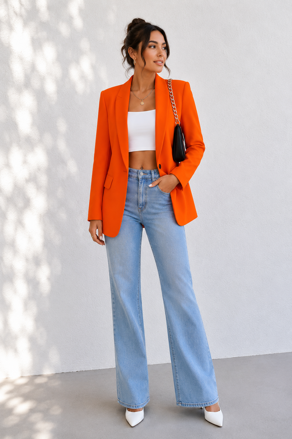 A woman stands against a light gray wall wearing a bright orange blazer over a white cropped tank top, light blue wide-leg jeans, and white pointed heels. She is holding a small black shoulder bag with a chain strap.
