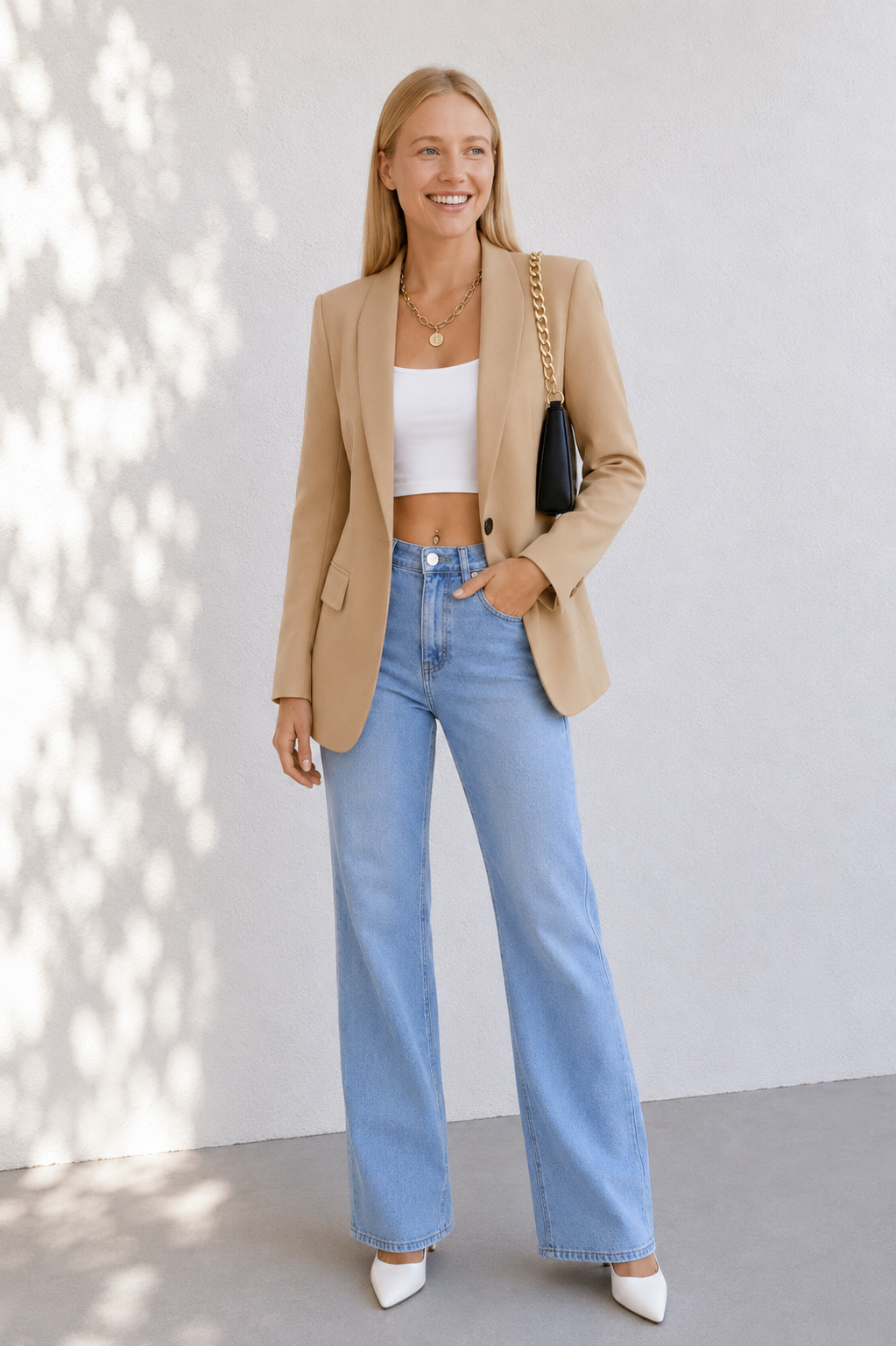 A woman stands against a light gray wall wearing a tan blazer, white crop top, light blue wide-leg jeans, and white pointed shoes, while carrying a black shoulder bag.
