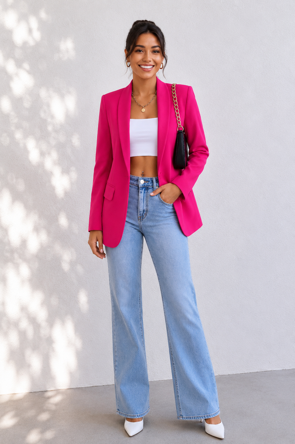 A woman modeling a bright pink blazer over a white crop top, paired with light blue wide-leg jeans, white pointed shoes, and carrying a black shoulder bag, set against a light gray wall with soft leaf shadows.