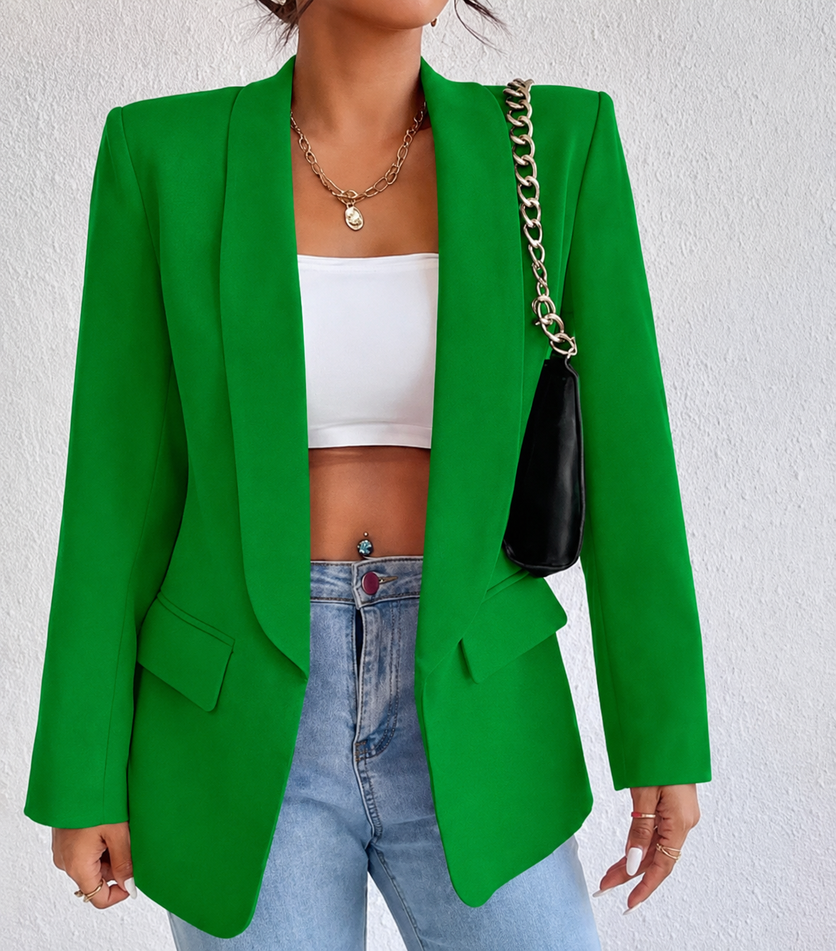 A woman wearing a bright green blazer over a white crop top and light blue jeans, carrying a black shoulder bag with a gold chain strap against a light textured wall.