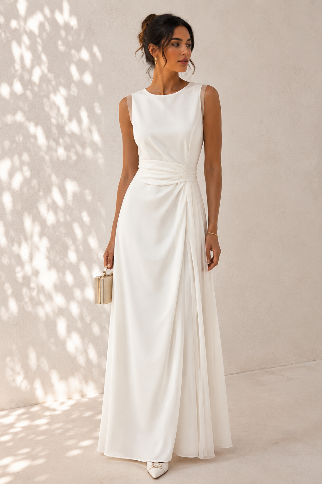 A woman models a sleeveless white floor-length gown with a gathered waist and draped skirt, standing against a light neutral wall with dappled sunlight and shadows.