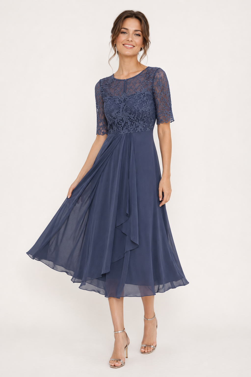 A woman modeling a navy blue midi dress with a lace bodice, sheer short sleeves, and a flowing chiffon skirt, paired with nude high-heeled sandals against a plain white studio background.
