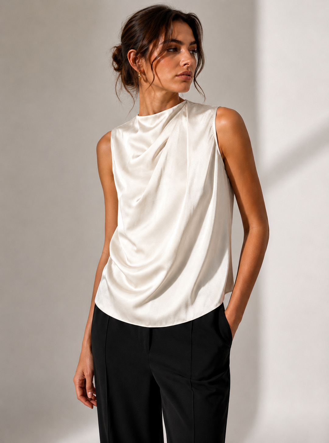 A woman wearing a sleeveless draped satin top in off-white and black trousers, standing against a neutral light gray wall with soft shadows.