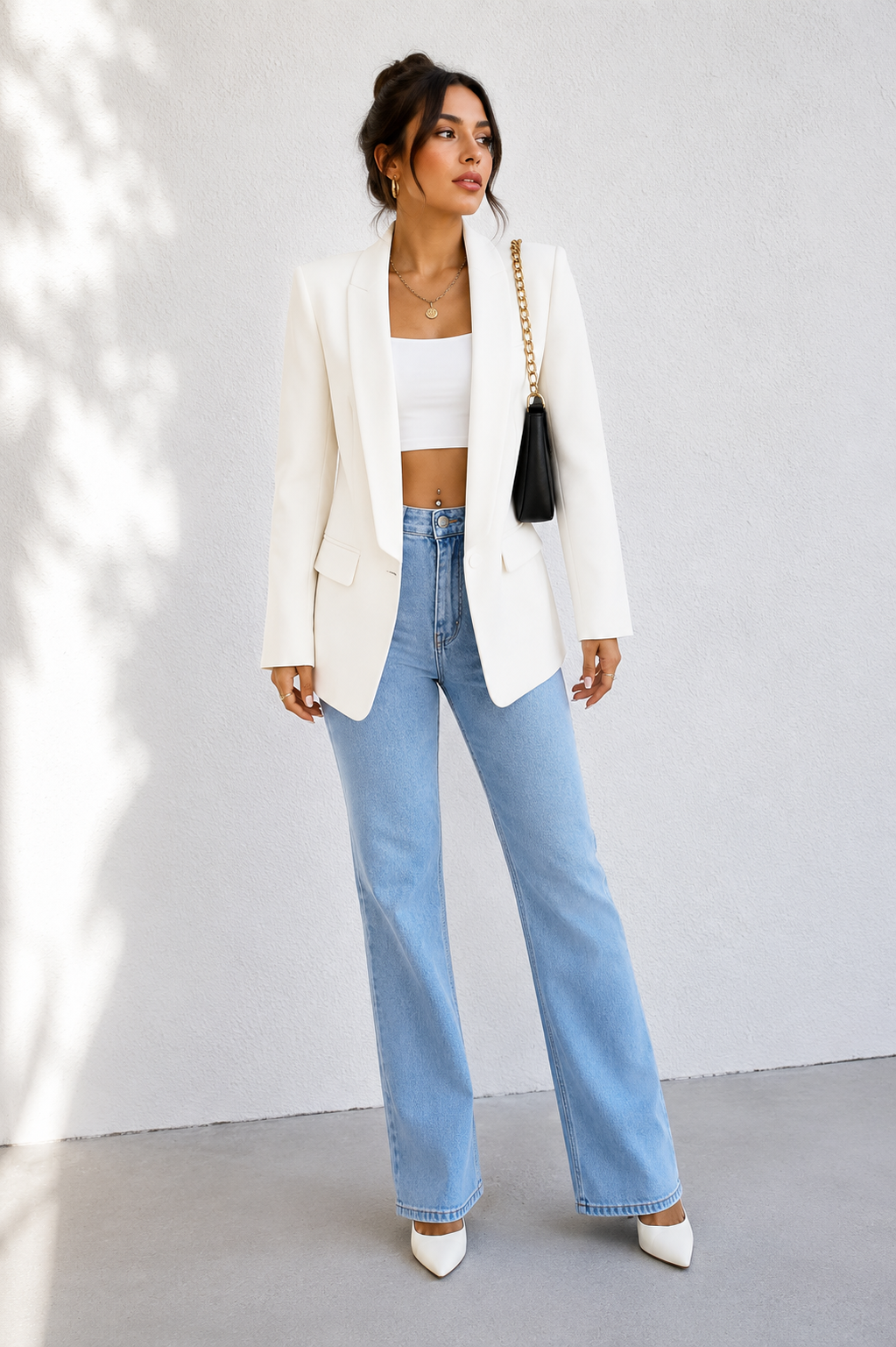 A woman models a white blazer, light-wash blue flare jeans, and white pointed-toe heels against a light gray wall with soft leaf shadows.