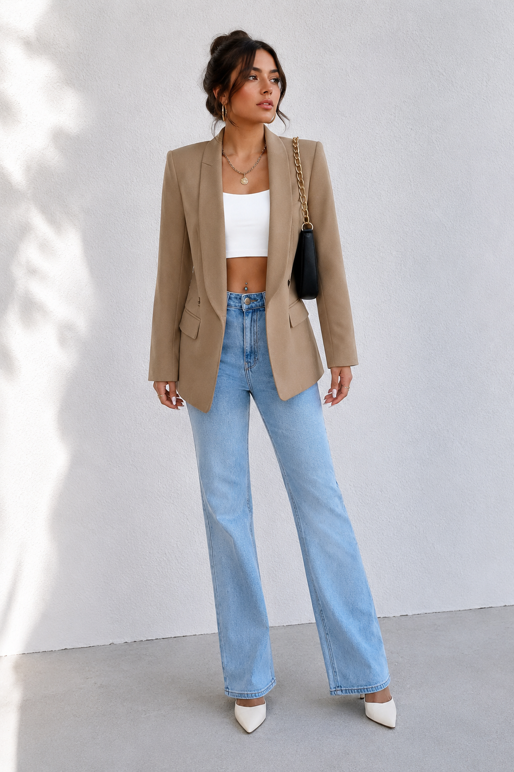 A woman stands against a light gray wall wearing a tan blazer over a white crop top, light blue wide-leg jeans, and white pointed heels, with a black shoulder bag.