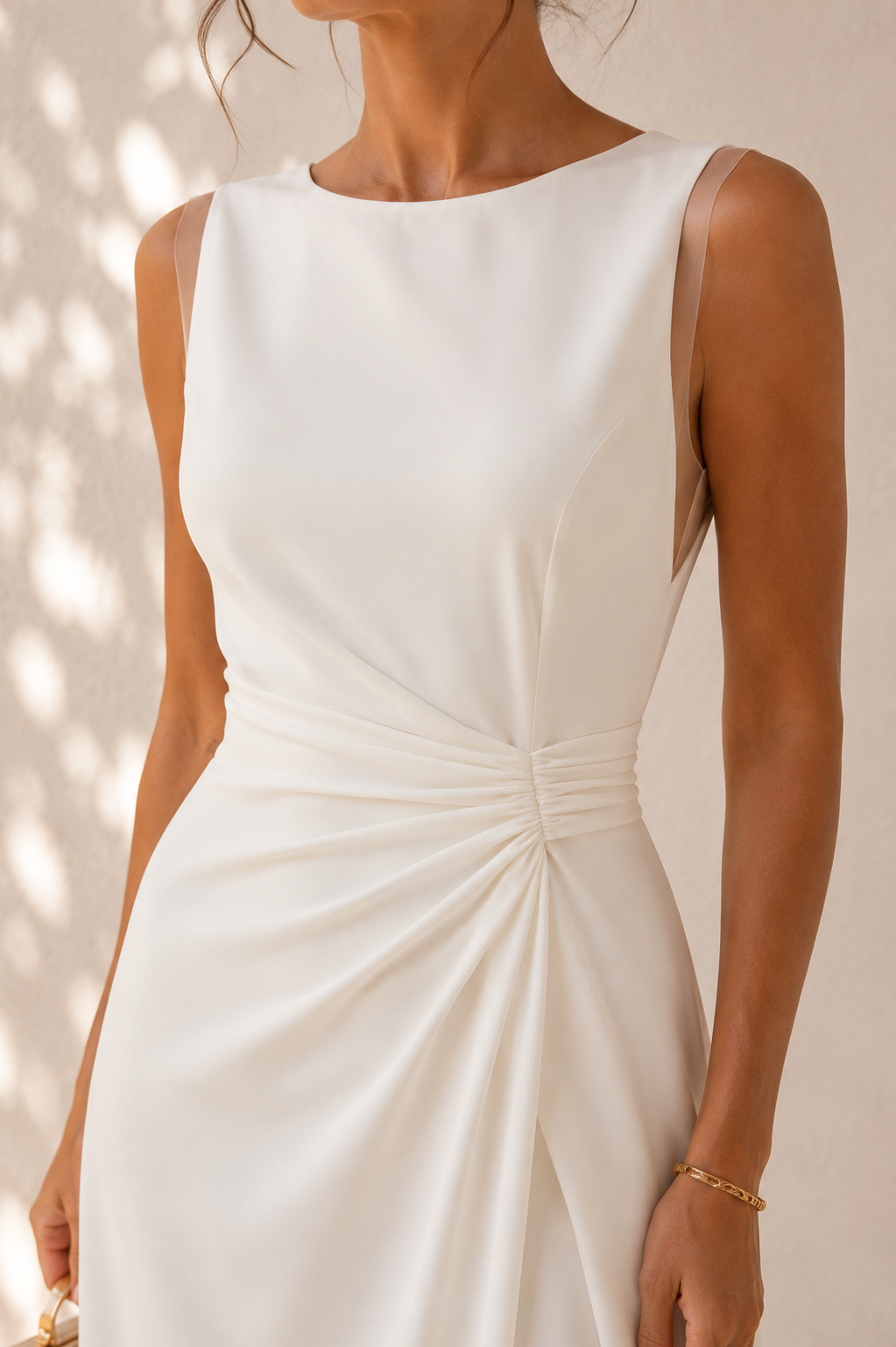 A woman wearing a sleeveless white dress with a boat neckline and a gathered ruched detail at the waist, shown against a softly lit neutral background.