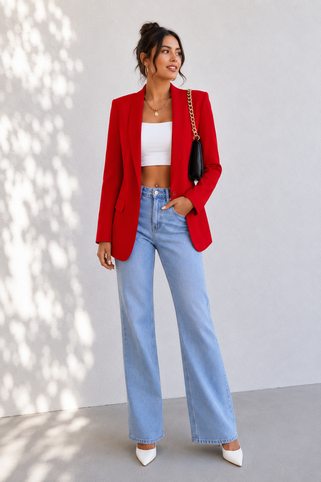 A woman stands against a light gray wall wearing a bright red blazer over a white crop top, light blue wide-leg jeans, and white pointed heels. She is carrying a black shoulder bag with a gold chain strap.