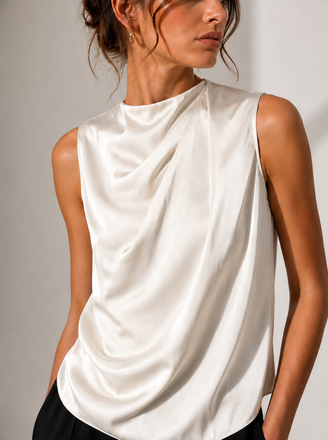 A close-up of a model wearing a sleeveless, high-neck top made of a soft, draped satin-like fabric in an off-white color.
